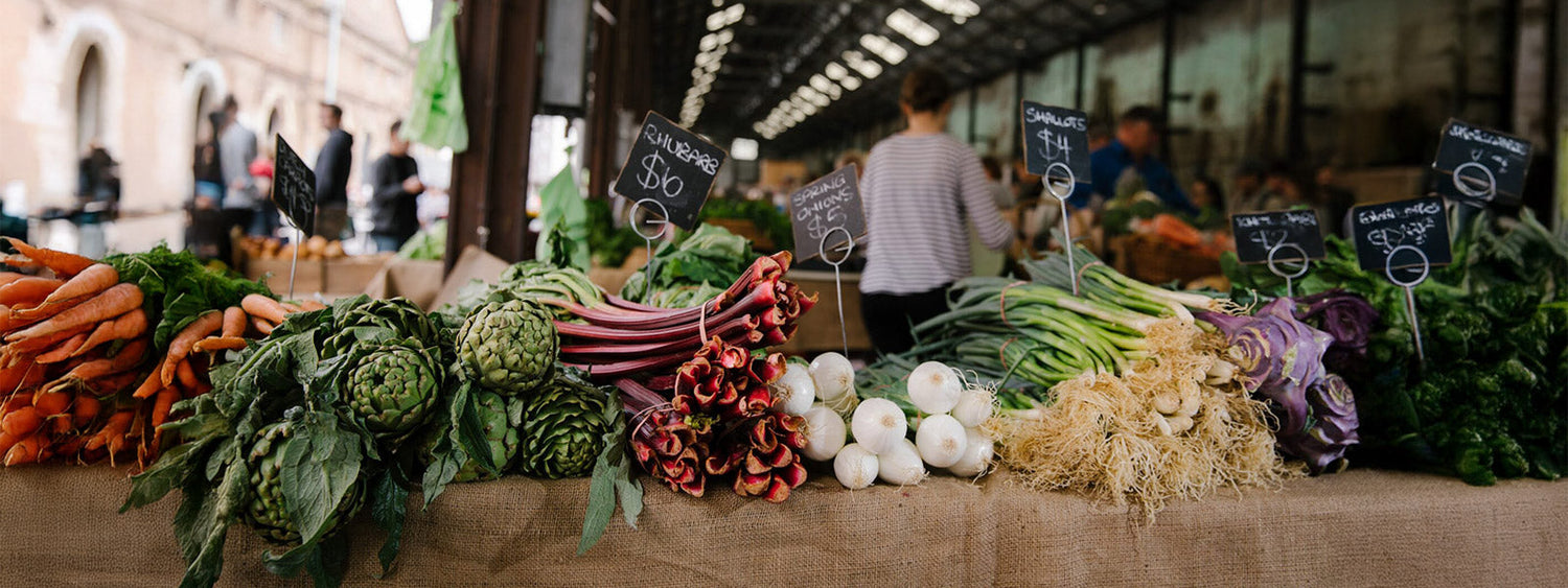 Fresh organic produce on display at Carriageworks Farmers Market in Sydney, including carrots, artichokes, rhubarb, spring onions, and shallots on a rustic stall.