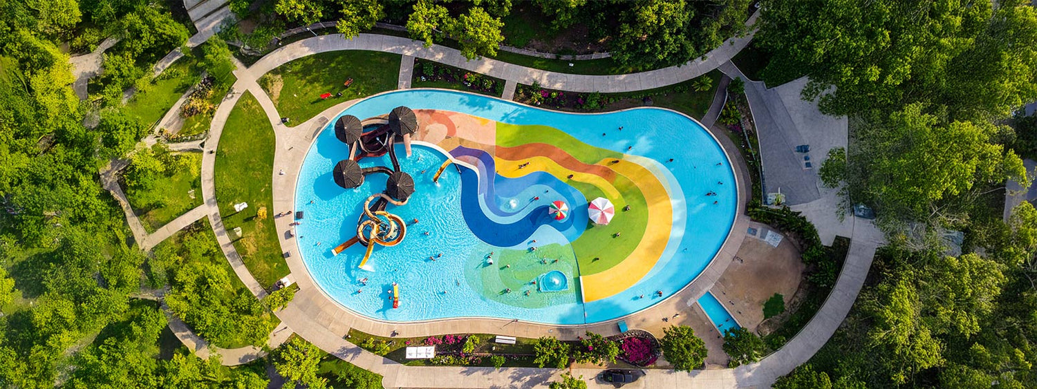 Aerial view of a colourful outdoor splash park surrounded by green lawns and walking paths on a sunny day, featuring fountains, slides and shallow pools.