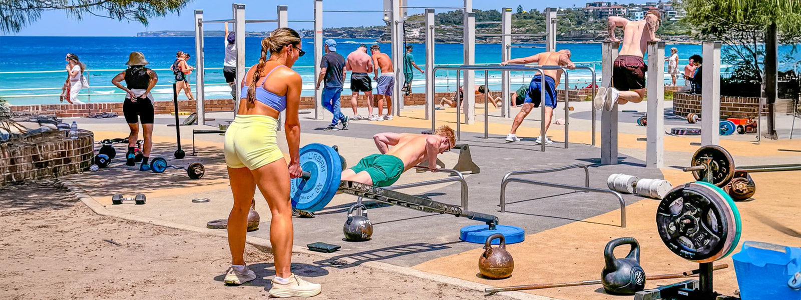 Outdoor gym at Bondi Beach with people weight training and kettlebell lifting on the sand under blue skies