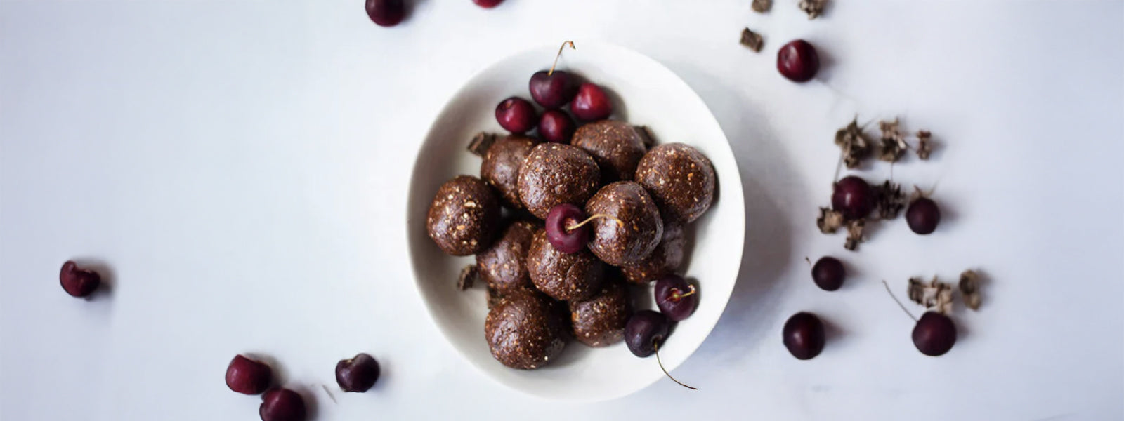 Bowl of no-bake chocolate cherry energy balls topped with fresh cherries, surrounded by scattered cherries on a white background.