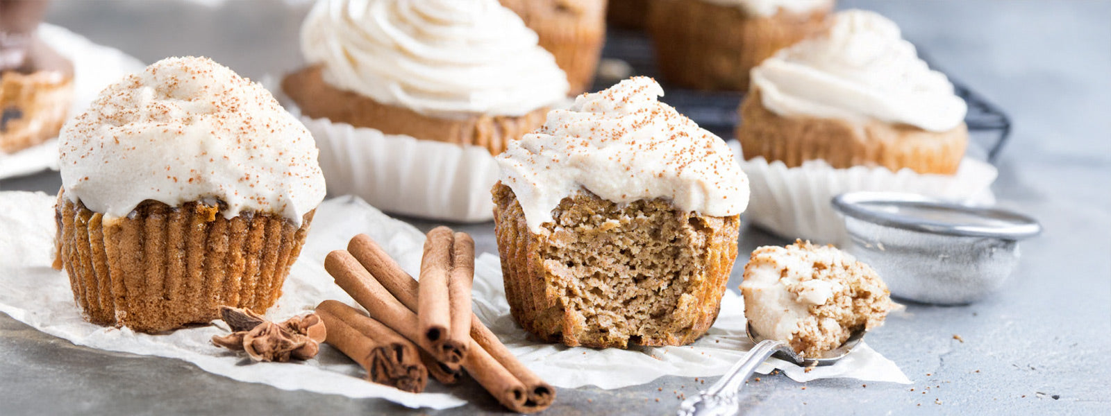 Paleo pumpkin cupcakes topped with creamy maple cashew frosting, one with a bite taken out, surrounded by cinnamon sticks and spices.