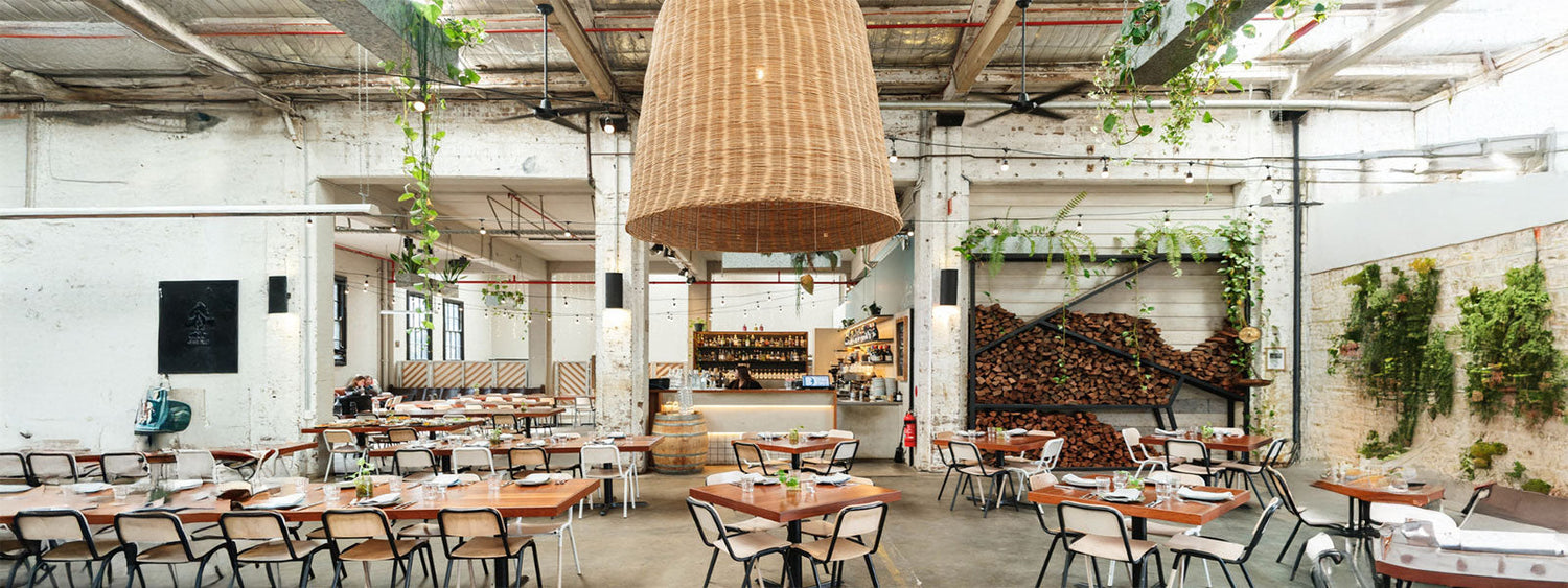 Interior of Three Blue Ducks Rosebery, a converted warehouse space featuring exposed brick walls, hanging plants, stacked firewood, industrial beams, and wooden tables set for dining beneath a large woven pendant light.